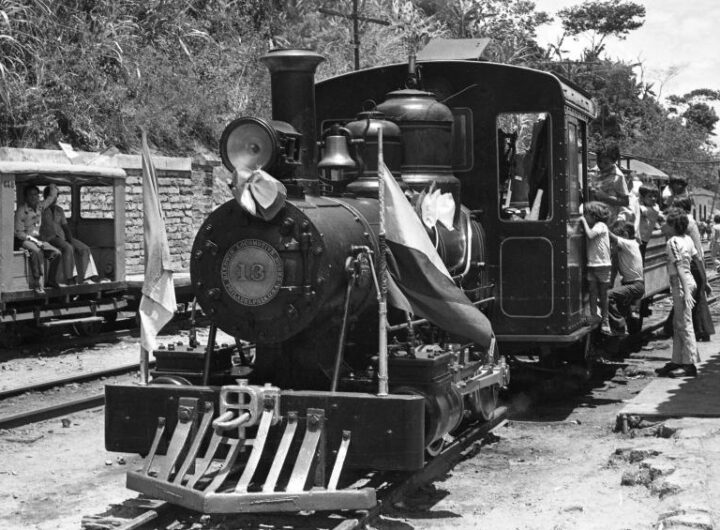 Locomotora histórica recorriendo una línea férrea entre montañas colombianas, símbolo del desarrollo ferroviario nacional.